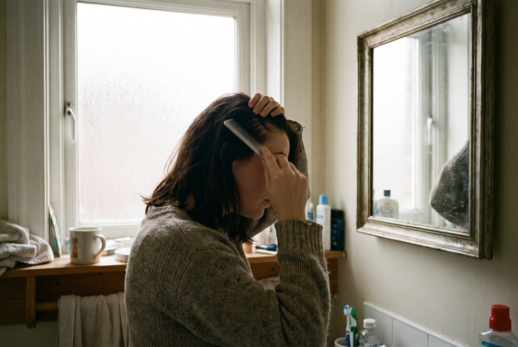 A woman in her twenties in a bathroom mirror reflection examining her hairline with a comb in hand