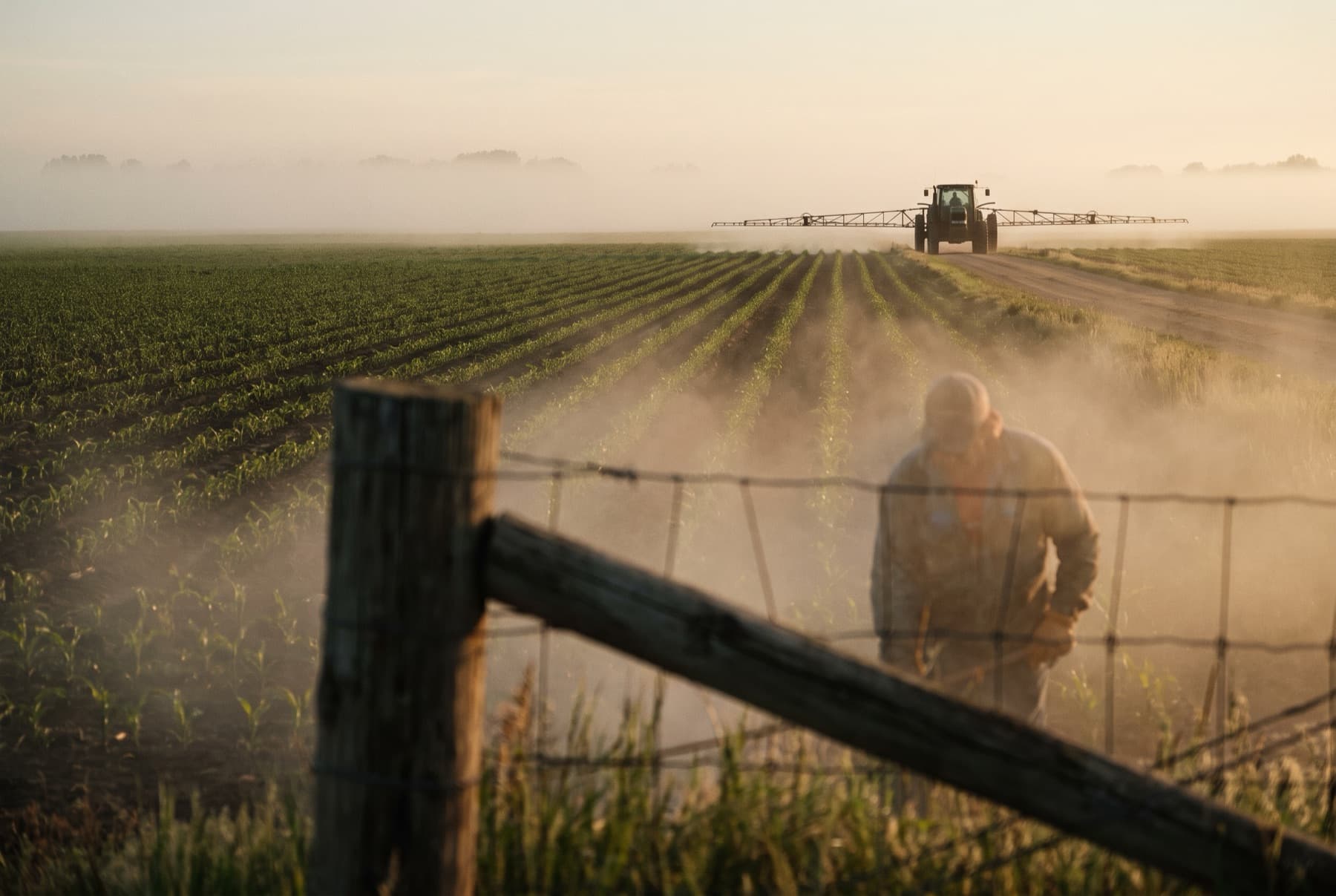 A farm field at sunrise with a tractor pulling a crop sprayer in the distance